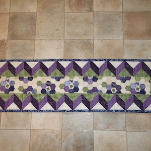 May include: A purple, green, and white quilted table runner with a geometric pattern. The runner features a central band of hexagonal shapes with floral designs, framed by a border of alternating purple and green triangles.