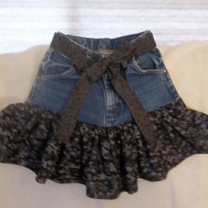 K&ouml;nnte beinhalten: Ein Jeansrock mit einem schwarzen Band, das um die Taille gebunden ist. Der Rock hat unten einen schwarzen und grauen R&uuml;schenbesatz.