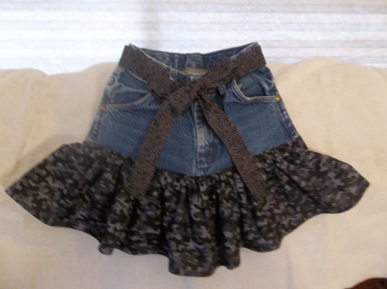 K&ouml;nnte beinhalten: Ein Jeansrock mit einem schwarzen Band, das um die Taille gebunden ist. Der Rock hat unten einen schwarzen und grauen R&uuml;schenbesatz.