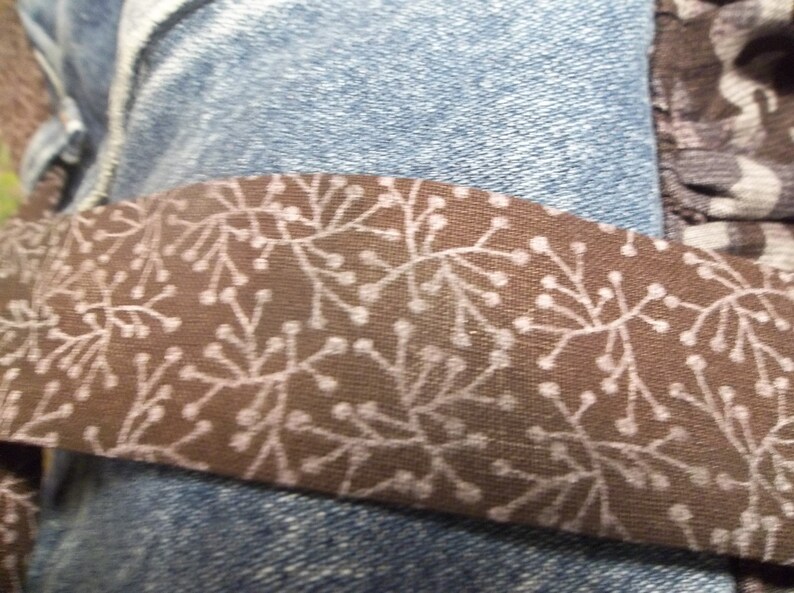 K&ouml;nnte beinhalten: Ein braunes Stoffband mit einem wei&szlig;en Blumenmuster. Das Band ist an einer blauen Jeans befestigt.