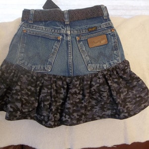 K&ouml;nnte beinhalten: Ein Jeansrock mit einem R&uuml;schen-Saum in Tarnmuster. Der Rock hat einen braunen Lederg&uuml;rtel mit einer silbernen Schnalle und ein Wrangler-Logo-Patch auf der Ges&auml;&szlig;tasche.