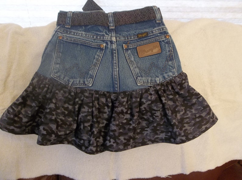 K&ouml;nnte beinhalten: Ein Jeansrock mit einem R&uuml;schen-Saum in Tarnmuster. Der Rock hat einen braunen Lederg&uuml;rtel mit einer silbernen Schnalle und ein Wrangler-Logo-Patch auf der Ges&auml;&szlig;tasche.