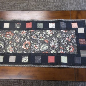 Puede incluir: Un camino de mesa floral blanco y negro con un borde negro y cuadrados rojos, verdes y blancos. El camino de mesa está sobre una mesa de madera.