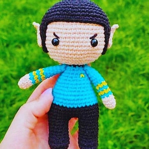 Könnte beinhalten: Handgemachte Häkelpuppe einer Figur mit schwarzem Topfschnitt, spitzen Ohren und einer blauen Uniform mit gelben Akzenten. Die Puppe hat eine schwarze Hose und wird vor einem verschwommenen grünen Hintergrund gehalten.