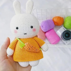 May include: A handmade white crochet bunny with black eyes, pink cheeks, and a small black stitched mouth. The bunny wears an orange dress with a carrot-shaped bag. The bunny is holding a carrot-shaped bag. The bunny is a cute, handmade toy.