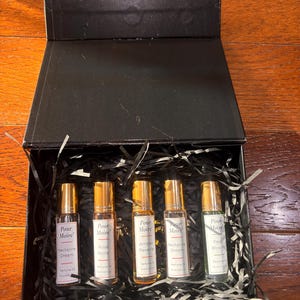 May include: A black gift box with five perfume oil bottles. Each bottle has a gold cap and a label with the brand name "Pour Moire" and a fragrance name. The fragrances are "Nectarine Dream", "Rose Noire", "Amber Reverie", "Nectarine Dream", and "Pear Bloom".