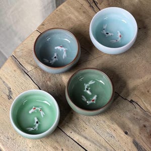 Puede incluir: Cuatro cuencos de cerámica en tonos verdes y azules, cada uno con un diseño de pez koi en 3D. Los cuencos tienen un acabado brillante y están dispuestos sobre una superficie de madera. Los peces koi tienen detalles en negro y rojo.