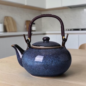 May include: A blue ceramic teapot with a dark brown woven handle and a matching lid. The teapot has a glossy finish and a spout on the left side. The handle is attached to the teapot with gold-colored metal pieces. The teapot is sitting on a light-colored wooden surface.