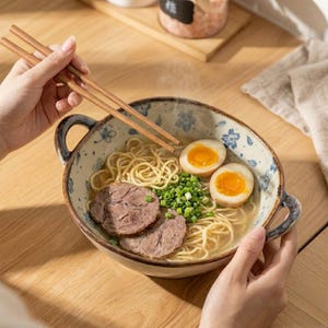 May include: A bowl of ramen with noodles, sliced meat, soft-boiled eggs, and green onions. The bowl has a floral design and is held with chopsticks. The ramen is a Japanese cuisine.