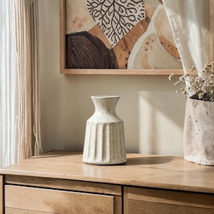 Könnte beinhalten: Eine cremefarbene, gerippte Keramikvase mit schmalem Hals steht auf einer Holzoberfläche. Die Vase ist der Mittelpunkt, mit einer neutralen Farbpalette. Ein gerahmtes Kunstwerk und eine Jutevase mit Trockenblumen befinden sich im Hintergrund.