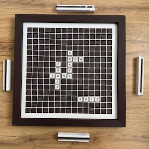 May include: A framed Scrabble board displaying the words "CRAVSTONE" and "EDIZ". The board has a brown grid with white tiles, set in a dark brown frame. Four tile racks are positioned around the frame.