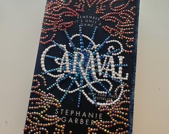 May include: A hardback book titled "Caraval" by Stephanie Garber. The cover is dark blue with the title in white, and the cover is decorated with colourful beads. The text "Remember it's only a game" is also visible.