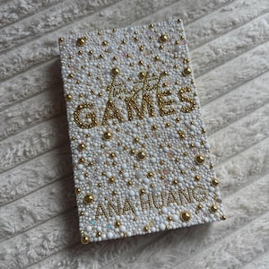 May include: A rectangular, white, bedazzled item with the words "The GAMES" and "ANA HUANG" in gold lettering. The surface is covered in small white and gold beads, with larger gold beads scattered throughout.