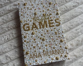May include: A rectangular, white, bedazzled item with the words "The GAMES" and "ANA HUANG" in gold lettering. The surface is covered in small white and gold beads, with larger gold beads scattered throughout.