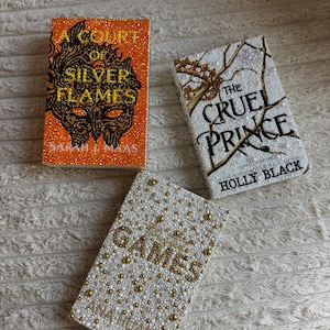 May include: Three hardback books with beaded covers. The titles are "A Court of Silver Flames", "The Cruel Prince", and "Games". The first book has an orange cover with black and gold beading. The second book has a white cover with gold beading. The third book has a white cover with gold beading.