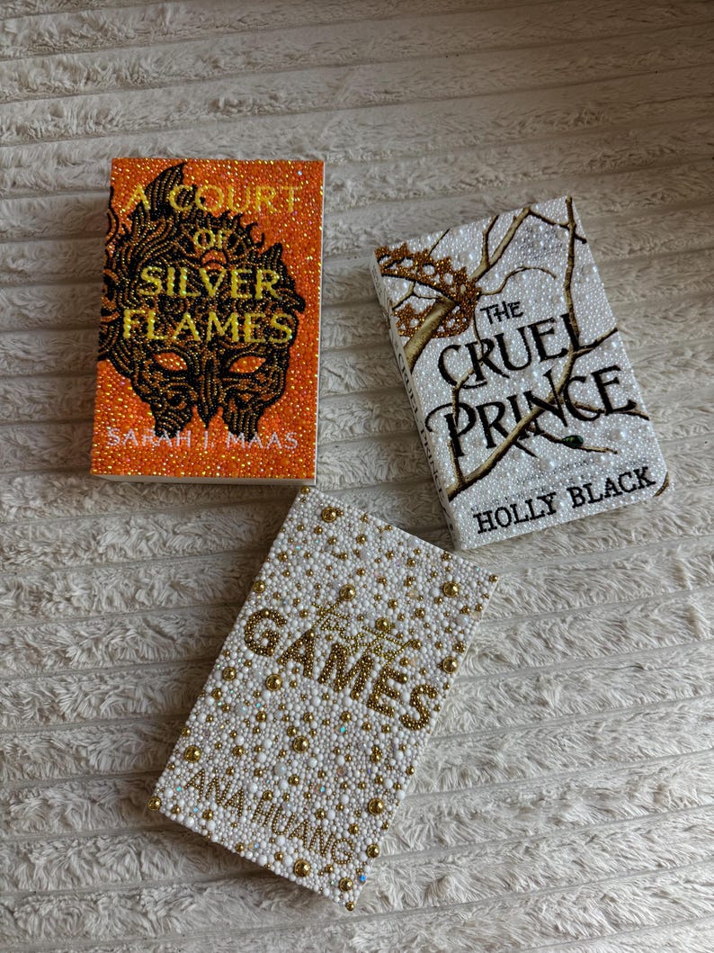May include: Three hardback books with beaded covers. The titles are "A Court of Silver Flames", "The Cruel Prince", and "Games". The first book has an orange cover with black and gold beading. The second book has a white cover with gold beading. The third book has a white cover with gold beading.