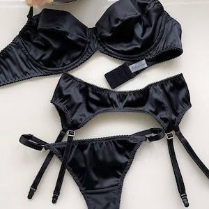 May include: Black satin lingerie set including a bra, garter belt, and thong. The bra has underwire support and adjustable straps. The garter belt and thong feature delicate trim. The set is displayed on a white surface.