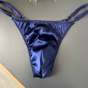 Puede incluir: Tanga de satén azul marino con delicadas correas y detalles de anillos metálicos plateados. La lencería presenta una textura suave y brillante y un diseño de tanga clásico. Los bordes están acabados con una costura decorativa.