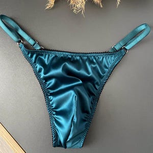 Könnte beinhalten: Ein String aus blaugrünem Satin mit glatter, glänzender Oberfläche und zarter schwarzer Bogenkante an den Rändern. Das Kleidungsstück verfügt über verstellbare, dünne Satinbänder, die vorne am Bund mit silberfarbenen Metallringen verbunden sind.