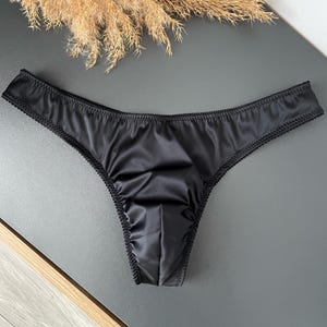 Op de afbeelding: Een zwarte satijnen string met een gladde, glanzende afwerking en een verfijnde zwarte picotrand langs de zomen. Het kledingstuk heeft een voorgevormd voorpand met subtiele rimpelingen bij de naden voor een gestructureerde pasvorm.
