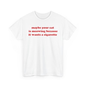Peut inclure: Un t-shirt blanc à manches courtes avec un texte rouge imprimé sur la poitrine qui dit, maybe your cat is meowing because it wants a cigarette. Le vêtement présente une encolure ronde et une coupe standard sur un fond blanc uni.