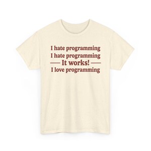 K&ouml;nnte beinhalten: Ein cremefarbenes Kurzarm-T-Shirt mit gesprenkelter Textur. Auf der Vorderseite befindet sich ein dunkelbrauner Text: I hate programming, I hate programming, &mdash; It works! &mdash;, I love programming. Das T-Shirt hat einen Rundhalsausschnitt und einen geraden Saum.