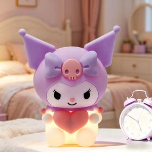May include: A light-up, cartoon-style night light in the shape of a character with a purple head, ears, and bow, and a pink heart. The night light has a smiling face and is sitting on a white surface. A pink alarm clock is next to the night light.