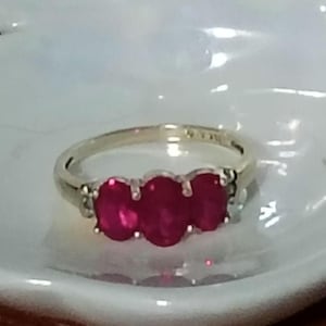 May include: A silver-toned ring featuring three oval-cut, deep red gemstones set horizontally in a row. The band is smooth and metallic, showcasing a classic three-stone design with prongs securing each vibrant red stone in place.