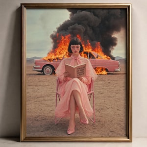 May include: A framed art print depicting a person with dark hair wearing a light pink dress and matching heels, sitting on a pink chair in a desert. Behind them, a pink car is engulfed in bright orange flames and thick black smoke. The person holds a book titled The Art of Not Giving a Fuck.