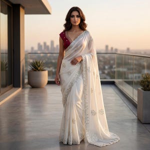May include: A white saree featuring silver embroidered floral motifs and a decorative border, paired with a deep maroon blouse with short sleeves and a V-neckline. The fabric is draped elegantly, showcasing intricate silver detailing throughout.