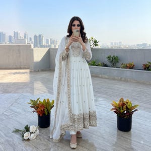 May include: A white floor-length Anarkali dress featuring intricate gold embroidery and mirror work on the bodice, sleeves, and hem. The outfit includes a matching sheer dupatta with a decorative gold border, paired with white trousers and flats.