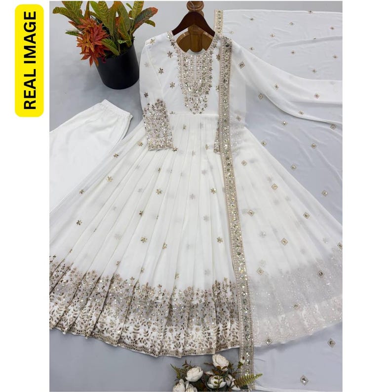 May include: A white long-sleeved floor-length dress featuring silver embroidery and mirror work on the bodice, cuffs, and hem. The dress includes a matching white dupatta with scattered silver embellishments and a pair of white trousers.