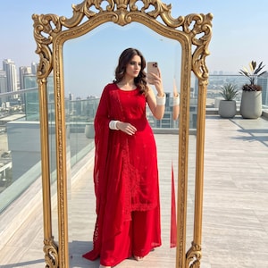 May include: A vibrant red sleeveless tunic paired with matching wide-leg trousers and a sheer red dupatta featuring intricate tonal embroidery. The ensemble is accessorized with white bangles on both wrists, creating a cohesive monochromatic look.