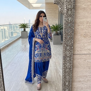 Könnte beinhalten: Ein königsblaues Salwar-Kameez-Set mit aufwendigen goldenen und bunten Stickereien mit Pfauenmotiven. Die langärmelige Tunika hat einen Rundhalsausschnitt mit Quasten, kombiniert mit einer passenden Plisseehose und einem transparenten Schal.