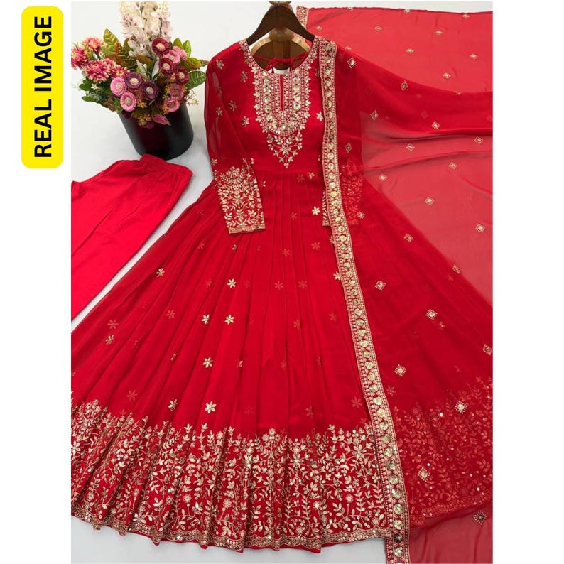 May include: A vibrant red floor-length gown featuring long sleeves, a round neckline with intricate gold embroidery and mirror work, a flared skirt with scattered floral motifs, a wide embellished hem, and a matching red dupatta and trousers.