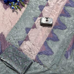 May include: A patterned textile in shades of pink, purple, and teal, with a scalloped edge. A small silver camera rests on the fabric, and a bouquet of blue flowers is placed nearby. The fabric has a shiny, silky appearance.