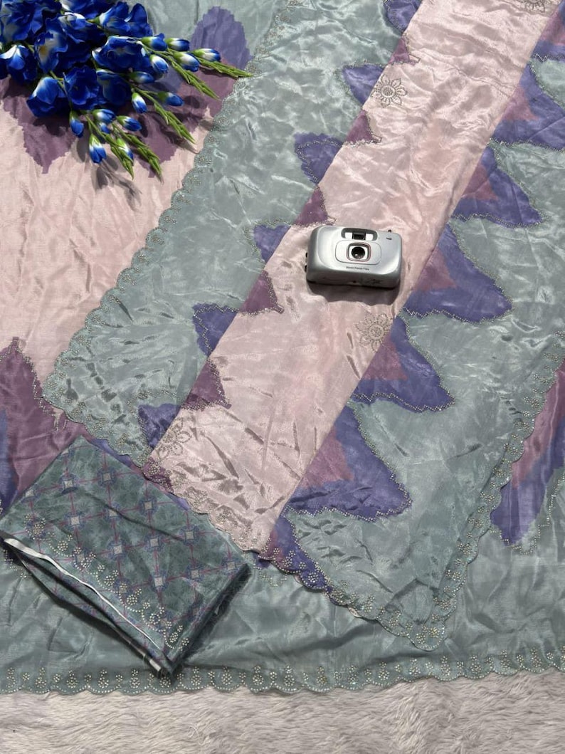 May include: A patterned textile in shades of pink, purple, and teal, with a scalloped edge. A small silver camera rests on the fabric, and a bouquet of blue flowers is placed nearby. The fabric has a shiny, silky appearance.