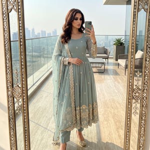 May include: A sage green long-sleeved Anarkali dress featuring a round neckline, a fitted bodice, and a flared skirt. The garment is adorned with intricate gold embroidery along the hem and cuffs, paired with a matching sheer dupatta and trousers.