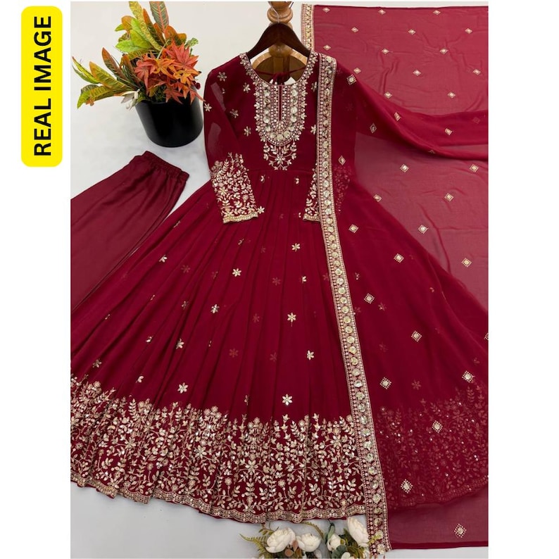 May include: A deep maroon long-sleeved floor-length dress featuring intricate gold embroidery around the neckline, cuffs, and hem. The dress includes a matching maroon dupatta with small gold accents and plain maroon trousers, all displayed on a hanger.