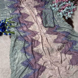 May include: A flowing, iridescent saree in shades of lavender, purple, and seafoam green. The fabric features a zigzag pattern with silver embellishments along the edges. A matching fabric piece is included. The saree is draped on a white surface.