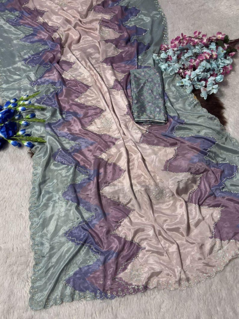 May include: A flowing, iridescent saree in shades of lavender, purple, and seafoam green. The fabric features a zigzag pattern with silver embellishments along the edges. A matching fabric piece is included. The saree is draped on a white surface.