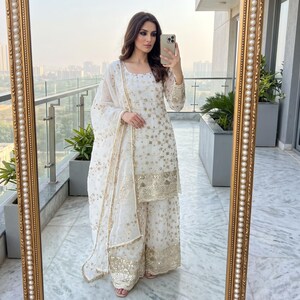 May include: A white three-piece outfit consisting of a long-sleeved tunic, wide-leg trousers, and a matching dupatta. The fabric features intricate gold embroidery and sequin embellishments throughout, with a wide decorative border at the hem.