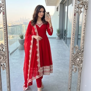 May include: A vibrant red long-sleeved dress featuring a flared skirt and matching slim-fit trousers. The ensemble is accented with intricate white and gold embroidery along the neckline, sleeve cuffs, hemline, and the border of a draped dupatta.