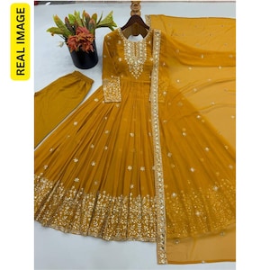 May include: A mustard yellow long-sleeved floor-length dress featuring intricate silver embroidery and sequin embellishments around the neckline, cuffs, and hem. The set includes matching mustard yellow trousers and a sheer, lightweight dupatta.