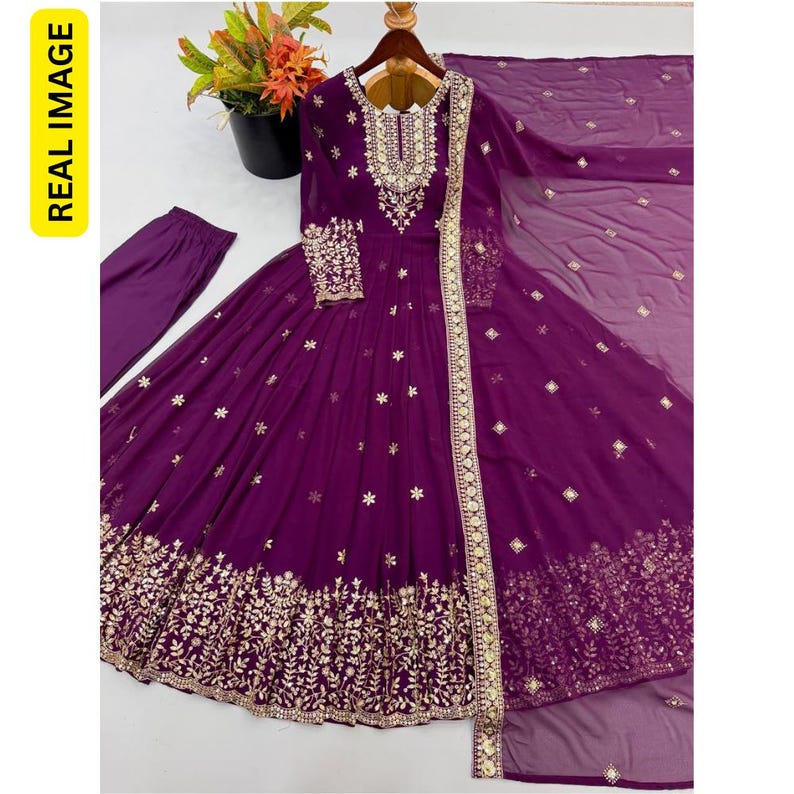 May include: A deep purple floor-length gown featuring intricate gold embroidery around the neckline, cuffs, and hem. The dress includes a matching sheer dupatta with gold accents and solid purple trousers, showcasing a floral and geometric pattern.