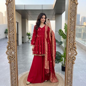 May include: A deep red peplum-style tunic featuring a gold floral pattern and small gold tassels along the hem, paired with a matching floor-length skirt and a long, gold-bordered red dupatta draped over the shoulder, captured in a large ornate mirror.