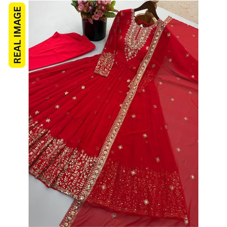 May include: A vibrant red long-sleeved dress featuring intricate gold embroidery and mirror work along the neckline, cuffs, and hem. The set includes matching red trousers and a sheer red dupatta with scattered gold floral embellishments.