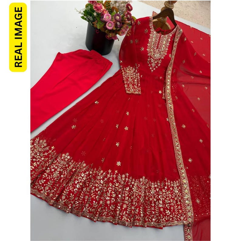 May include: A vibrant red long-sleeved floor-length dress featuring intricate gold embroidery and mirror work along the neckline, cuffs, and hem. The set includes matching red trousers and a sheer red dupatta with gold decorative borders.