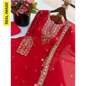 May include: A vibrant red long-sleeved dress featuring intricate gold embroidery and reflective mirror embellishments around the neckline and cuffs. The garment includes a matching sheer red dupatta with a decorative border and small floral motifs.
