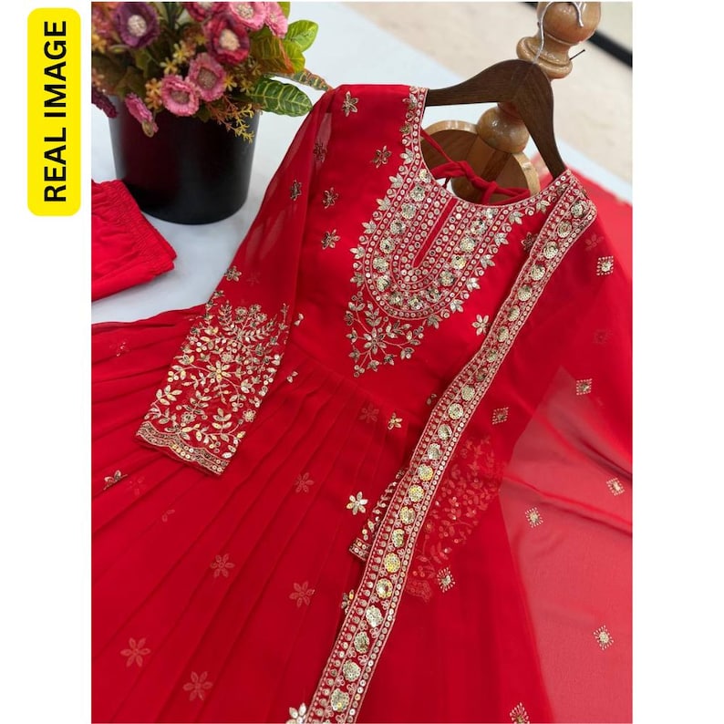 May include: A vibrant red long-sleeved dress featuring intricate gold embroidery and reflective mirror embellishments around the neckline and cuffs. The garment includes a matching sheer red dupatta with a decorative border and small floral motifs.